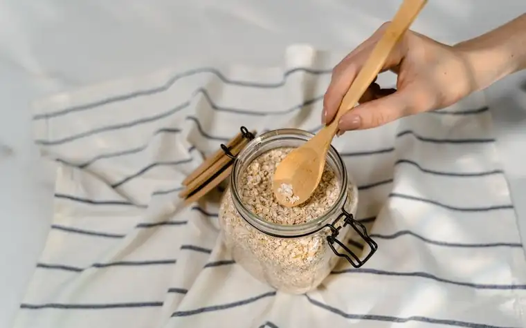 Hand scooping uncooked oats with a wooden spoon from a glass jar on a striped towel