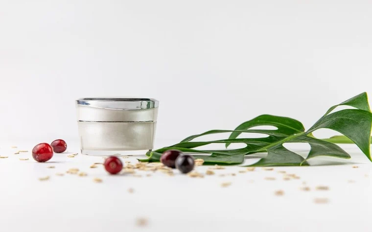 White cosmetic cream jar beside a green leaf, cranberries and scattered oats on a white surface