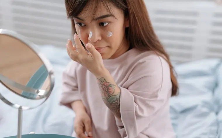 A woman applying cream to her face in front of a small mirror